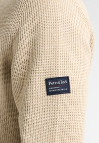 Beige stickad tröja med en texturerad design, med en mörkblå märkesetikett på ärmen som visar "Petrol Ind."