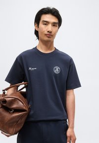 Άνδρας που φορά μπλε μπλουζάκι με σχέδιο και μπλε παντελόνι, κρατώντας μεγάλη καφέ δερμάτινη τσάντα σε απλό φωτεινό φόντο.