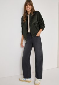 Mörkgrön bomberjacka med ribbad mudd, ihopparad med grå vida jeans. Vita sneakers med ljusa accenter. Släta tygtexturer synliga.