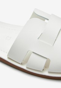 Sandalia deslizante de piel blanca con amplias correas recortadas, plantilla plana y suela de goma marrón. Textura suave con costuras precisas.