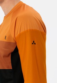Langarm-Sportshirt aus orangefarbenem und schwarzem Mesh-Gewebe; zeichnet sich durch atmungsaktiven Stoff, kontrastierende Farben und ein kleines Logo auf der Schulter aus.