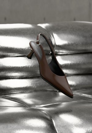 Chaussure à talon haut à bride arrière en cuir brun reposant sur une surface capitonnée métallique argentée brillante.