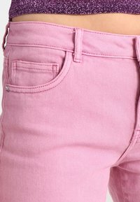 Jeans de denim rosa con una textura suave, diseño de cinco bolsillos y detalles cosidos. Presillas para el cinturón visibles y cierre de botón metálico.