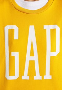 Maglietta di cotone gialla con grandi lettere bianche a blocchi che formano la scritta "GAP." Scollo rotondo con bordo bianco, texture liscia e senza motivi aggiuntivi.
