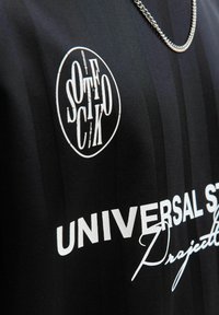 Maillot noir avec des rayures horizontales ; présente un logo blanc et un texte disant "UNIVERSAL STUDIOS" et "PROJECT." Le matériau semble lisse.