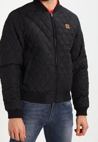 Černá prošívaná bomber bunda s zipovým uzávěrem, žebrovanými manžetami a čtvercovou koženou nášivkou na levém horním boku.