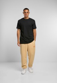 Melna kokvilnas T-krekls saskaņots ar bēšām, brīvā stilā šortiem ar oranžu logo. Balti sporta apavi papildina apģērbu.