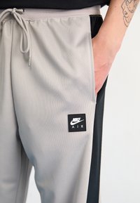 Grå joggingbyxor med en svart sidpanel, dragsko i midjan och en svart fyrkantig lapp med "NIKE AIR"-logotyp. Slät textur, atletisk design.