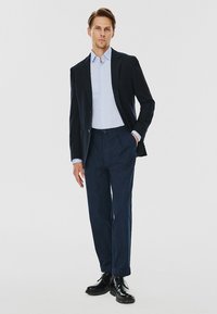 Granatowa marynarka o kroju dopasowanym noszona na jasnoniebieskiej koszuli, w zestawieniu z granatowymi spodniami. Czarne buty, gładka faktura, kroj dopasowany. Prosty design.