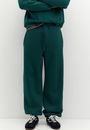 Personne portant un pantalon de survêtement ample vert foncé avec cordon de serrage et des baskets noires avec des lacets blancs, bras croisés au-dessus de la taille.