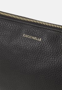 Bolso de mano de cuero negro con superficie texturizada, cremallera dorada y logotipo impreso que dice "COCCINELLE".