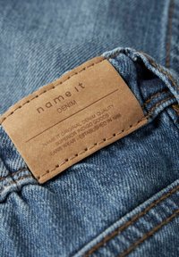 Traperice s smešnim kožnim etiketom na kojoj je utisnuti tekst: "nazovi to DENIM, ORIGINALNA KVALITETA DENIMA, osnovano 1986.", sa šivenim rubovima.