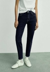 Vaqueros de talle alto de mezclilla oscura con corte ajustado, que incluyen el clásico estilo de cinco bolsillos y costuras amarillas, combinados con zapatillas blancas.