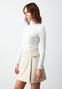 Witte geribde langecoat met lange mouwen, gecombineerd met een beige minirok met een gouden kettingaccent aan de zijkant en een gestructureerde pasvorm.