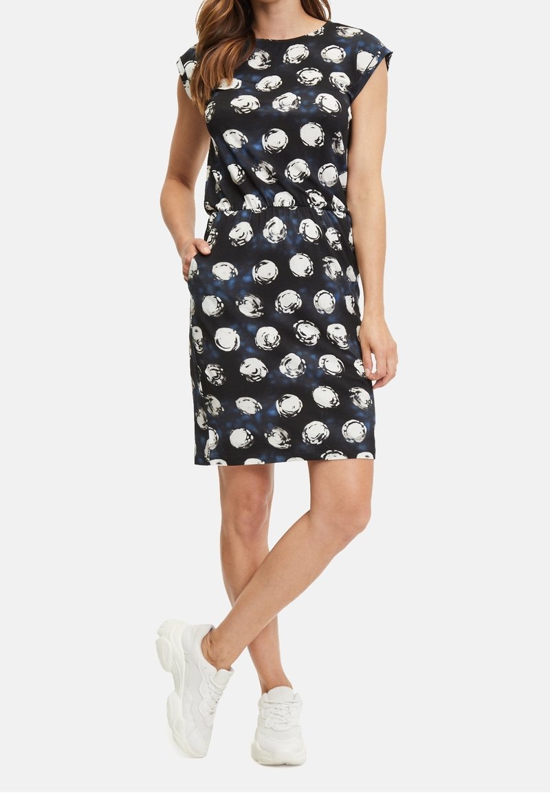 Robe noire à motifs circulaires blancs, manches courtes, taille élastique, poches et longueur genoux, associée à des baskets blanches.