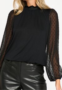 Blusa nera con maniche puff trasparenti testurizzate, colletto alto, tessuto arricciato e motivo a pois sulle maniche, abbinata a pantaloni neri lucidi.