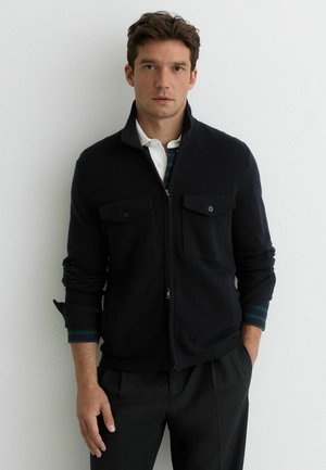 Mann trägt eine schwarze Jacke mit Reißverschluss und Klappentaschen über einem weißen Hemd mit Kragen und schwarzen Hosen, steht vor einer weißen Wand.