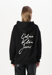 Felpa nera con cappuccio in cotone, con un logo bianco in corsivo sulla parte posteriore che dice "Calvin Klein Jeans". Vestibilità ampia con orlo a coste.