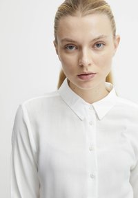 Blouse blanche à boutons avec un col et des manches longues, en tissu léger, présentant une texture lisse et un design classique.