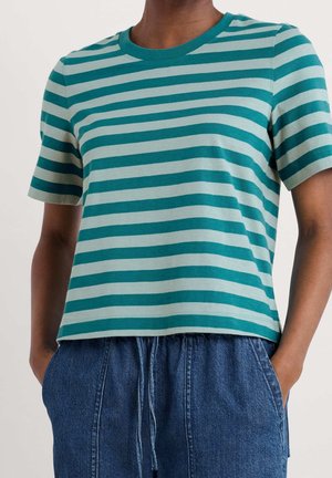Persoon draagt een teal en lichtblauw horizontaal gestreept T-shirt met korte mouwen en losse blauwe spijkerbroek met handen in de zakken.