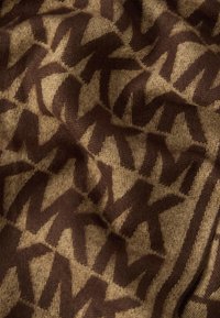 Barna és bézs színű textil, amely ismétlődő, egymásba fonódó "M" alakzatok mintáját ábrázolja. Puha textúrájú, kötött hatású anyag.