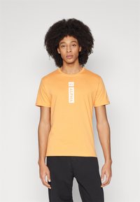 Orange bomulds t-shirt med en central vertikal hvid label med teksten "ALPHA." Korte ærmer og rund halsudskæring.
