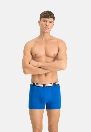 Oben-ohne-Mann steht mit verschränkten Armen da und trägt blaue Puma-Boxershorts vor einem schlichten hellen Hintergrund.