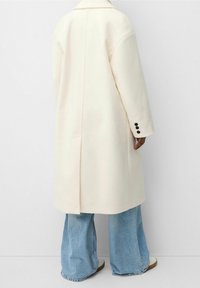 Witte mantel van een wolmix met een rechte pasvorm, grote revers en zwarte knoopdetails op de manchetten; gecombineerd met lichtblauwe jeans met wijde pijpen.