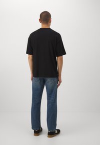 Camiseta negra de manga corta con un ajuste relajado, combinada conjeans rectos azules y zapatos negros, vista desde la parte trasera sobre un fondo liso.