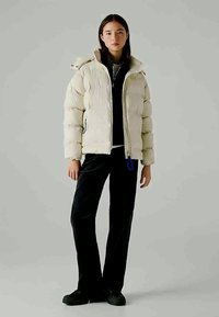 Pufferjack in lichtbeige, met een rits sluiting, capuchon en geribbelde manchetten. Draagt over een zwarte top met uitlopende zwarte broeken.