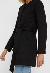 Black coat made of textured fabric, featuring a belted waist, long sleeves, and a minimalistic design with side pockets.