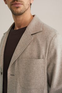 Helepruun tekstuuriga bleiser tipplapiga, ühe tasku ja mustade nuppudega, kantud tumepruuni särke peal.