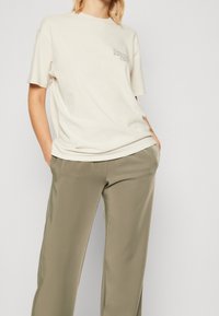 Crème-kleurige katoenen T-shirt met korte mouwen, voorzien van een kleine bedrukte graphic aan de linkerkant. In combinatie met olijfgroene, op maat gemaakte broek.