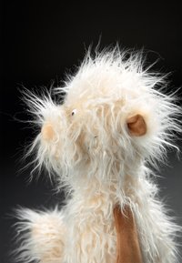 Fluffy cream and tan plush toy with a round muzzle, soft textured fur, small black eyes, and a brown ear, shown in profile against a dark background.