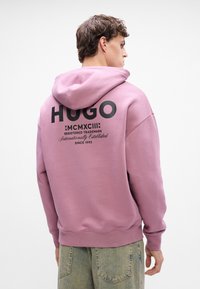 Hanorac roz cu un mare logo negru "HUGO" pe spate, având glugă cu șnur și manșete ribbate, purtat cu pantaloni din denim albastru deschis.