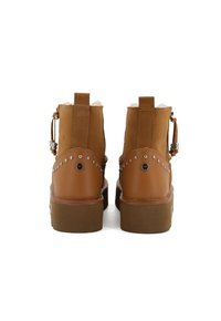 Para brązowych skórzanych i zamszowych botków za kostkę z metalowymi ćwiekami i futrzanym wykończeniem, widziana z tyłu na białym tle.
