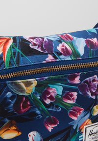 Tissu à motif floral avec des tulipes et d'autres fleurs aux couleurs vives, avec une fermeture éclair dorée et une étiquette blanche Herschel.