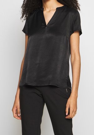 Mujer con blusa negra de manga corta y escote en V y pantalones negros sobre un fondo claro y liso.