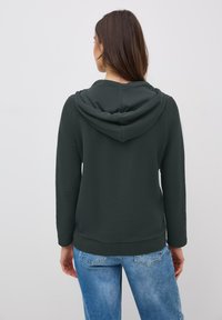 Felpa verde scuro con tessuto texturizzato, dotata di cappuccio e maniche lunghe, abbinata a jeans chiari strappati.