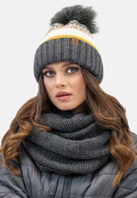 Grijze gebreide beanie met witte en gele strepen en een zwarte pompon, gecombineerd met een dikke grijze gebreide sjaal. Gelaagd stof, knusse ontwerp.
