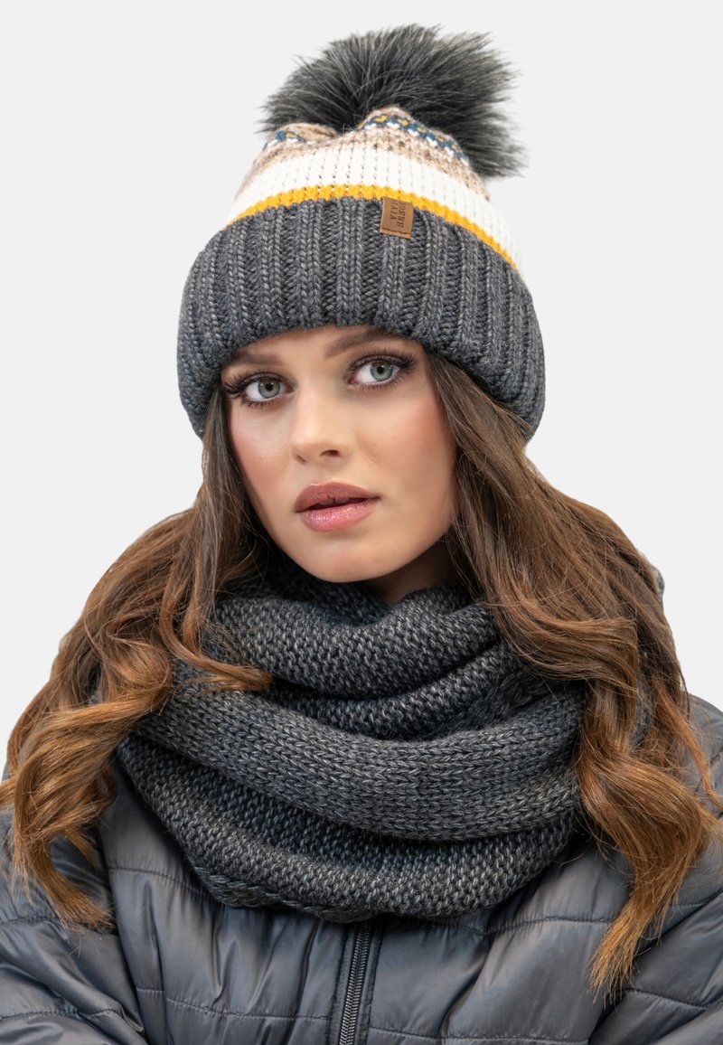 Grijze gebreide beanie met witte en gele strepen en een zwarte pompon, gecombineerd met een dikke grijze gebreide sjaal. Gelaagd stof, knusse ontwerp.