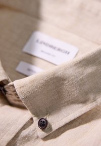 Cuello de camisa de lino beige con un botón marrón y una etiqueta que dice "Lindbergh, Talla M". Muestra una textura fina y detalles de tela ligera.