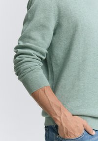Hellgrüner, gestrickter Pullover mit gerippten Bündchen. Der Stoff wirkt weich. Sichtbares Detail einer Hand in einer Tasche, die die Hautstruktur zeigt.