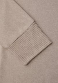 Mouw van een beige gebreide trui met geribbelde manchetdetails op een bijpassende beige stoffen achtergrond.