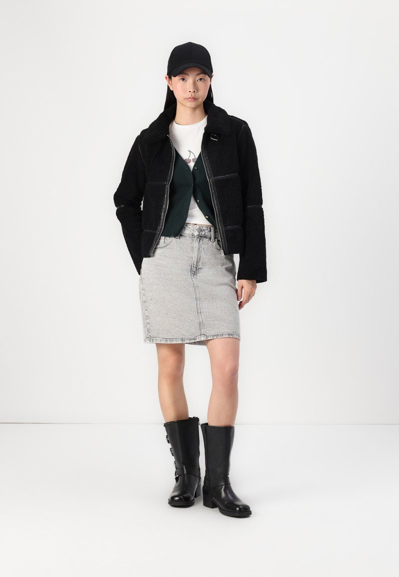 Giacca in pile nera con colletto, gilet verde sopra una maglietta bianca con grafica, gonna di jeans grigio chiaro e stivali neri al ginocchio con fibbie.