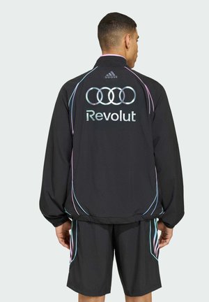 Mężczyzna odwrócony tyłem, ubrany w czarną kurtkę i szorty Adidas z neonkowym wykończeniem, na plecach kurtki znajduje się logo Audi i napis "Revolut".