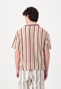 Cămașă cu mâneci scurte, cu guler, de culoare crem cu dungi roșii, dintr-un material tricotat texturat, cu o margine ușor curbată, asortată cu pantaloni cu dungi.