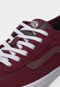 Κοντινό πλάνο μπορντό σουέντ αθλητικού παπουτσιού με λευκά κορδόνια, λευκή ραμμένη λωρίδα στο πλάι και λογότυπο Vans Off The Wall σε μαύρη ετικέτα στη γλώσσα.