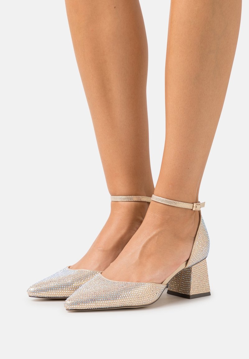 Femme portant des chaussures à bout pointu, talon bloc avec brides à la cheville, recouvertes de petits strass brillants sur un fond neutre.