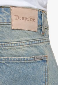 Gros plan d'un jean en denim bleu clair avec un patch en cuir et une étiquette en métal affichant tous deux le nom de la marque Dropsize.